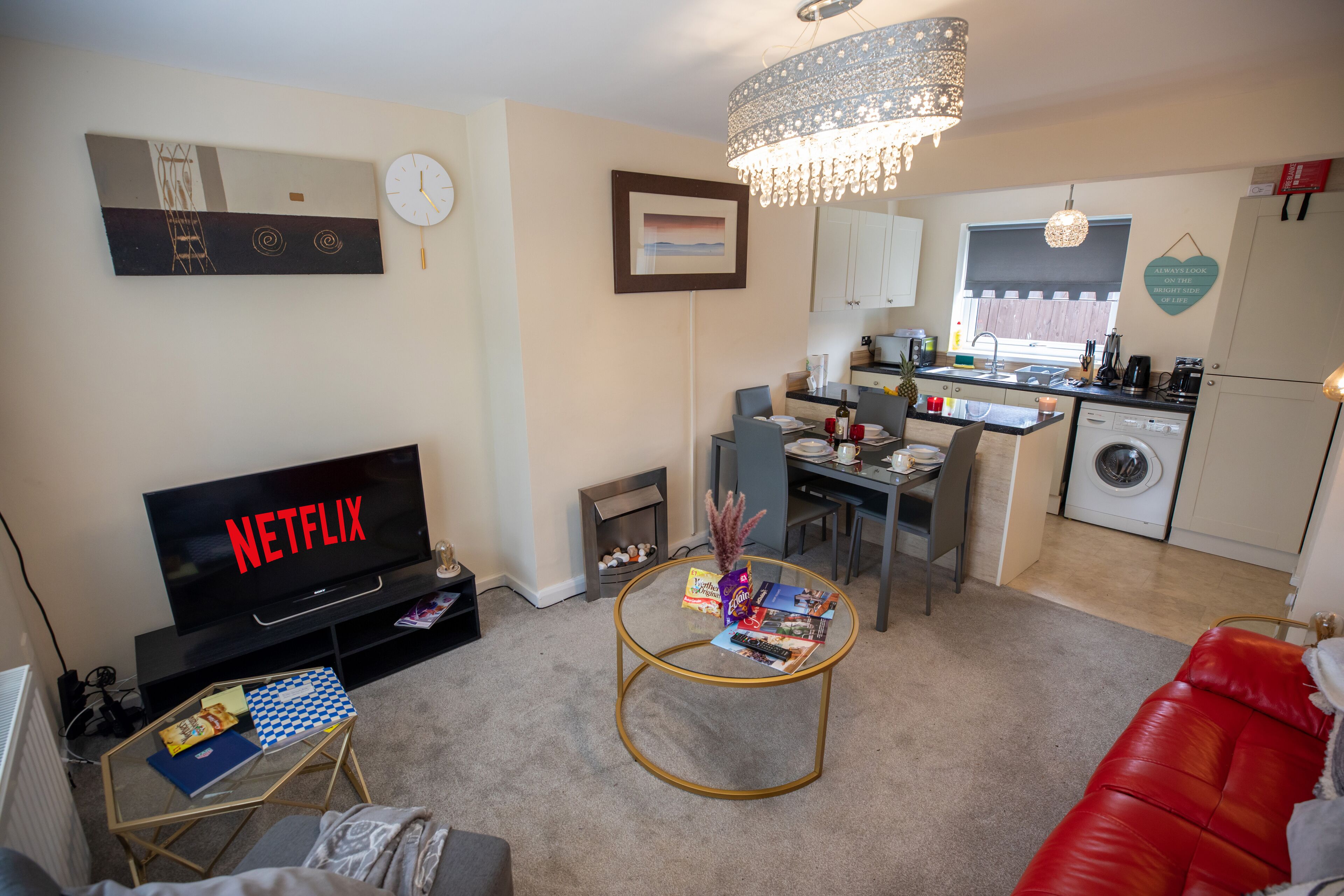 A modern living room with a flat-screen TV, a coffee table with magazines, a red sofa, and a kitchen area with a washing machine.