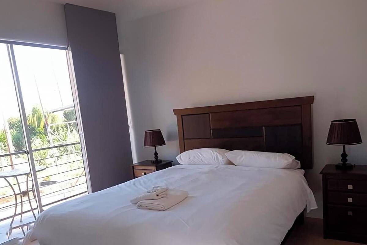Une chambre d’hôtel avec un grand lit, deux tables de chevet avec des lampes, une fenêtre donnant sur un paysage verdoyant et un balcon.