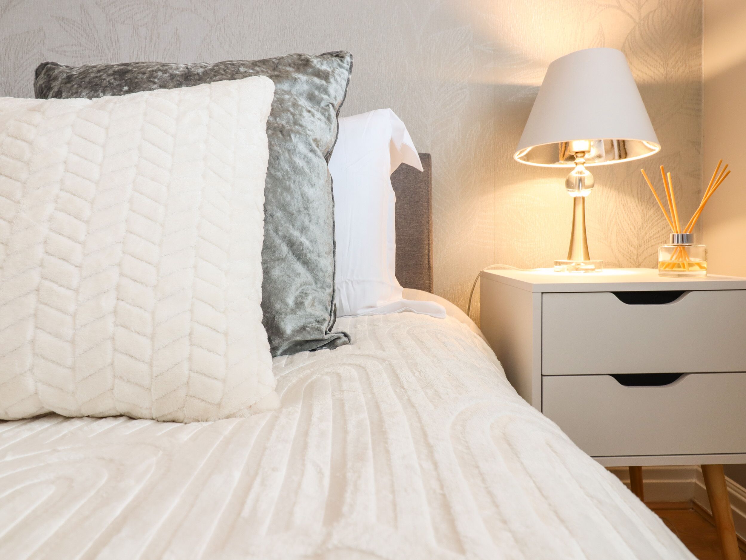 A neatly made bed with white and grey pillows, a white nightstand with a lamp and reed diffuser, and a wall with a subtle leaf pattern.