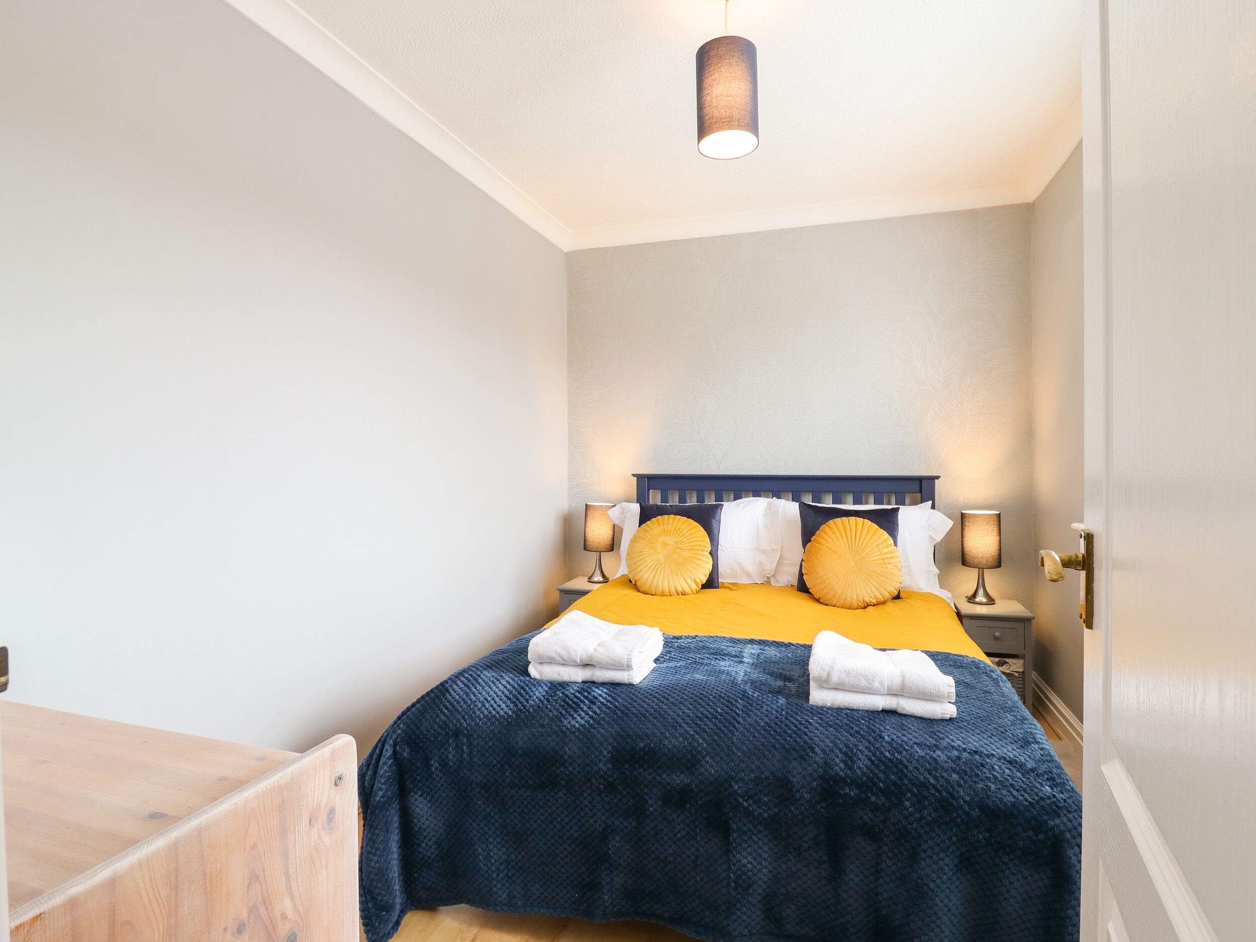 Een net opgemaakt bed met een blauw-gele sprei, twee sierkussens en twee bedlampjes.