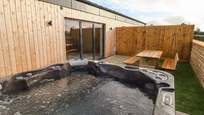 A hot tub in a wooden deck area with a sliding glass door and a picnic table. - 1 Bears Court Little Rissington (Cheltenham)