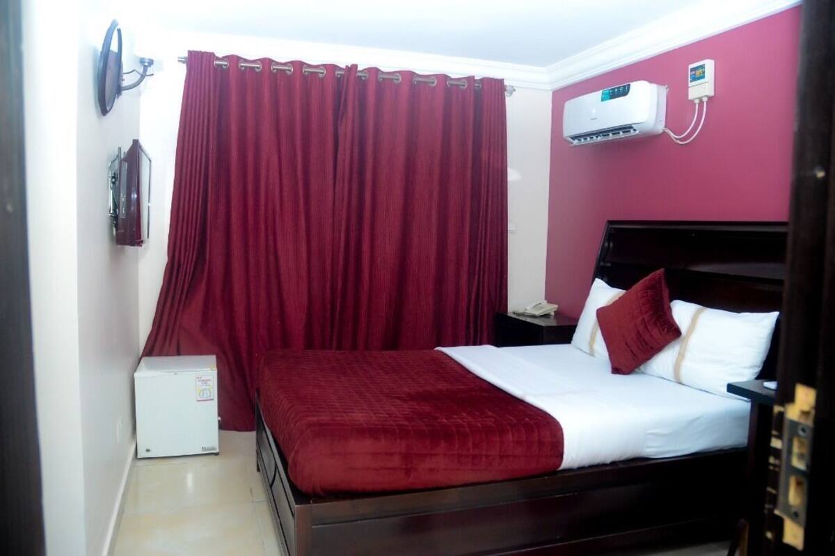 A hotel room with a bed, a nightstand, a wall-mounted air conditioner, and a red curtain.