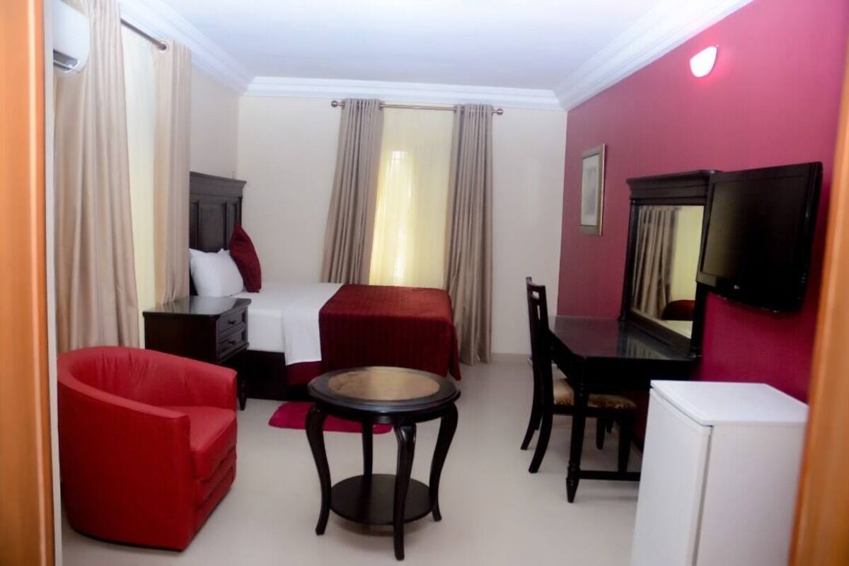 A hotel room with a bed, a red armchair, a small round table, a TV, and a white refrigerator.