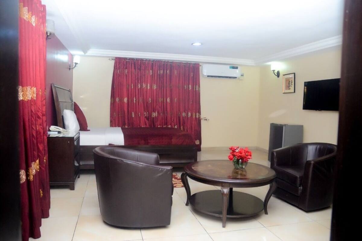 A hotel room with a bed, a sofa, two armchairs, a small round table with a vase of flowers, a TV, and an air conditioning unit on the wall.