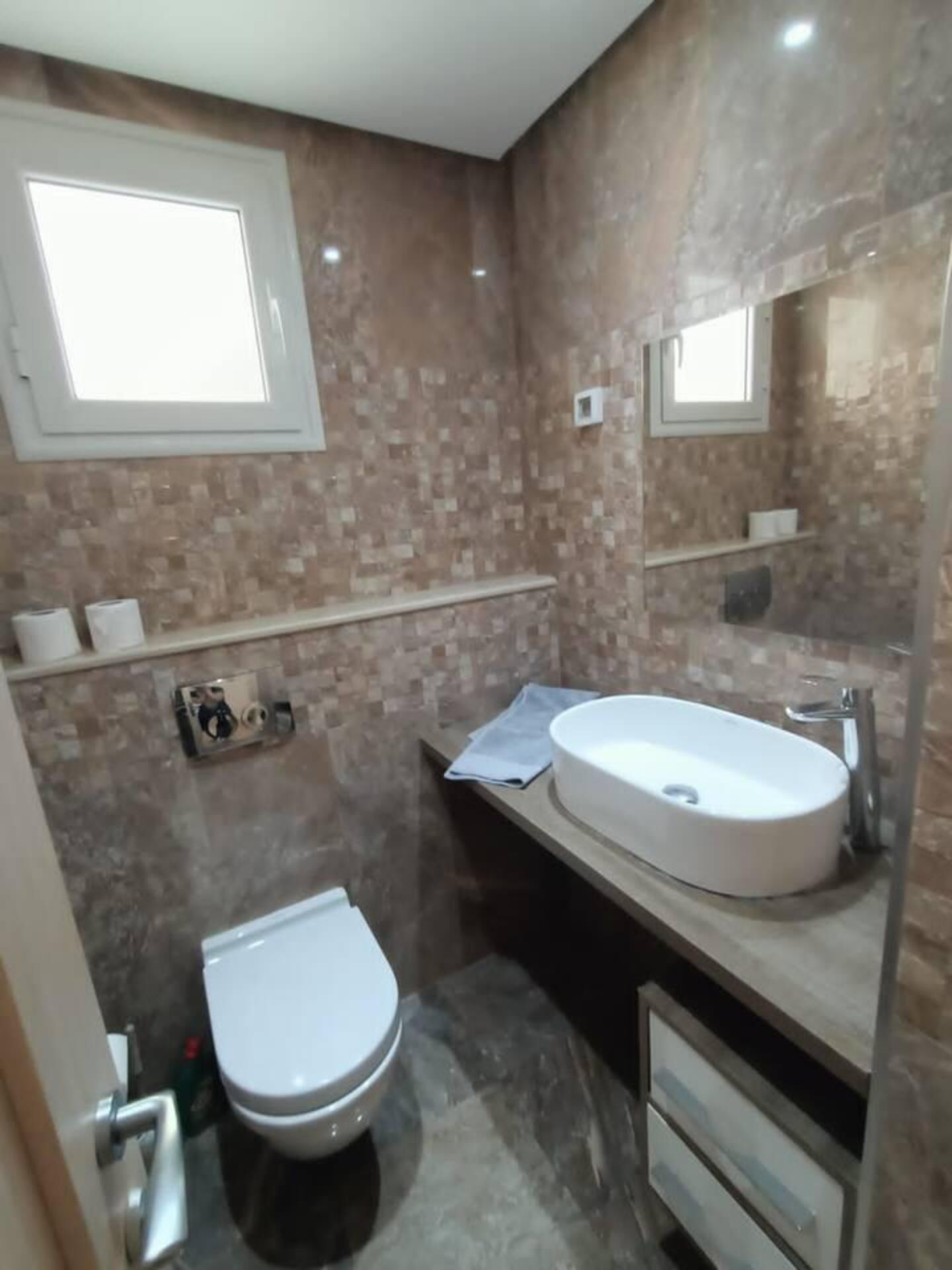 A bathroom with a toilet, sink, and mirror. There is a window above the toilet and a shelf with toiletries.