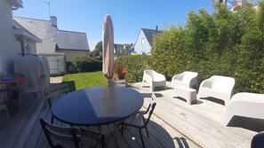 Um terraço com uma mesa redonda e quatro cadeiras brancas, ladeado por um deck de madeira e um viveiro.