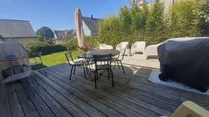 An outdoor deck with a round table and chairs, surrounded by greenery and a barbecue grill. - Maison au Calme Près des Plages (Concarneau)