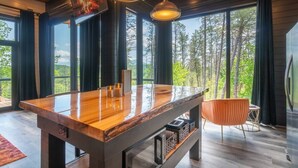 Una habitación amplia con una gran mesa de madera, dos lámparas colgantes y vistas al bosque a través de ventanas amplias.