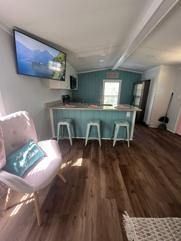 Une cuisine moderne avec des meubles de couleur turquoise, un grand îlot central doté de tabourets, un téléviseur à écran plat fixé au-dessus de l’îlot et une fenêtre laissant entrer la lumière naturelle.