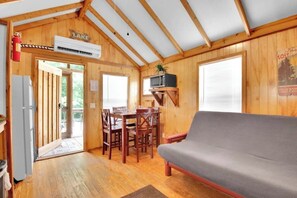 Une cabane avec un intérieur en bois, un coin repas avec une table et des chaises, un canapé gris et un téléviseur fixé au mur.