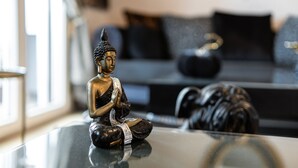 Une statue de Bouddha est posée sur une table en verre dans un salon moderne, avec un canapé et des coussins en arrière-plan.