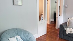 Een woonkamer met een grijze fauteuil, een grijze bank, houten vloeren en een witte deur die naar een andere kamer leidt.