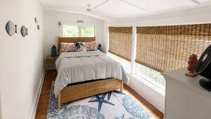 Een slaapkamer met een bed, een houten hoofdbord, een nachtkastje, een bijzettafeltje, een raam met jaloezieën en een ventilator bevestigd aan de muur.