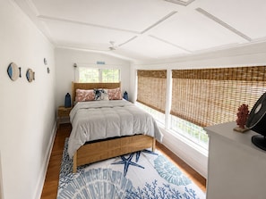 Un dormitorio con una cama, cabecera de madera, mesita de noche y persianas en la ventana. Incluye un ventilador de pared.