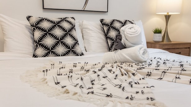 Un lit bien fait, recouvert d’une couverture à motifs, avec deux oreillers à dessins noir et blanc et une lampe de chevet.