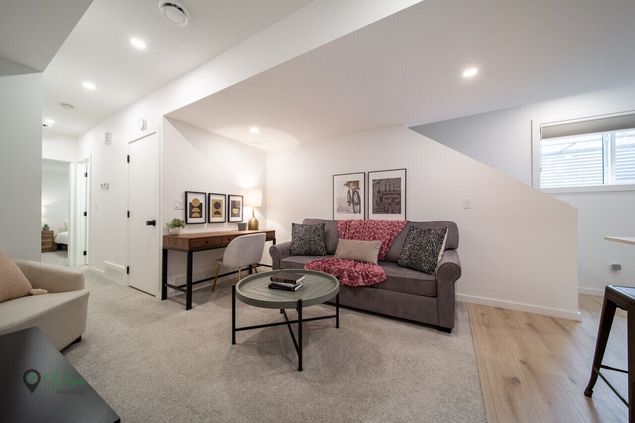 A modern living room with a grey sofa, a round coffee table, and a desk with a chair. There are framed pictures on the wall and a window with blinds.