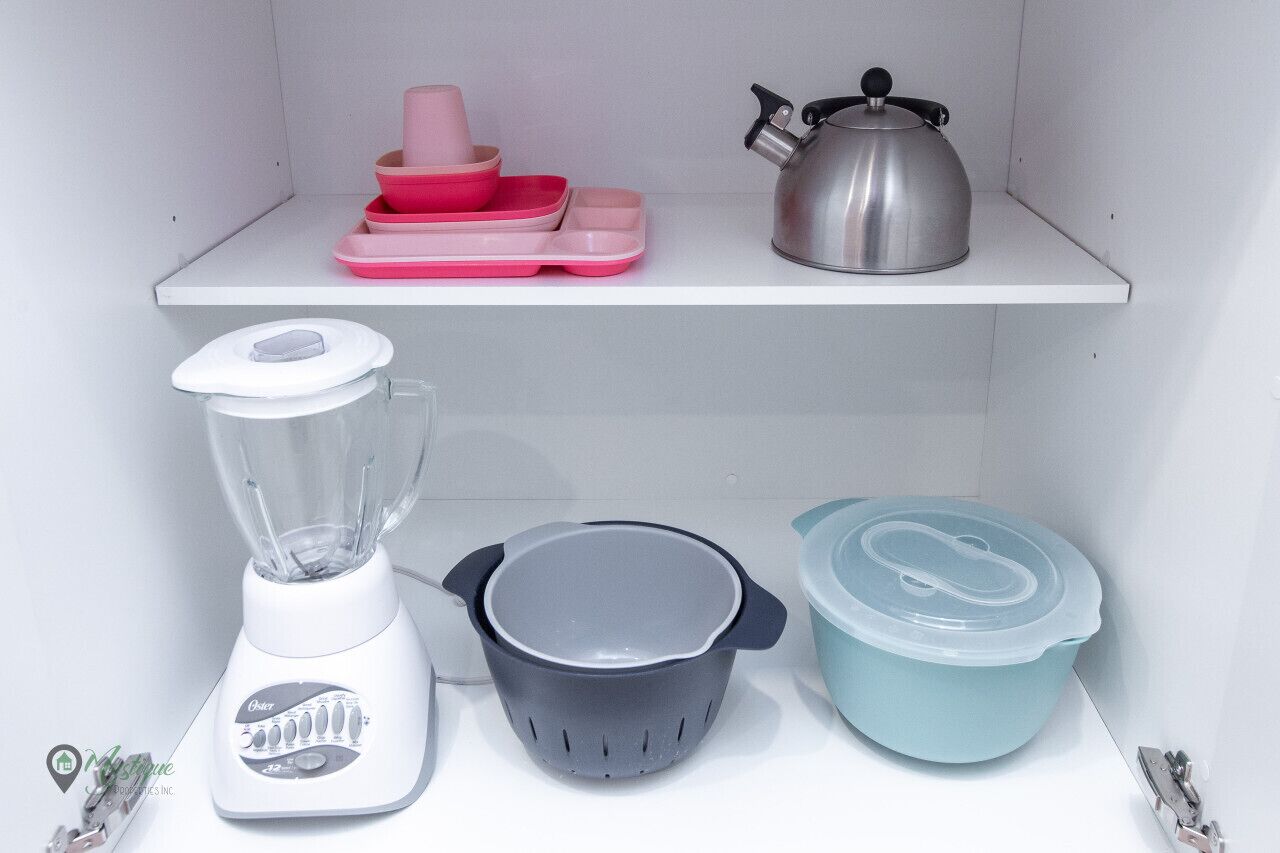 A kitchen shelf with a blender, a kettle, a colander, and pink and blue containers.