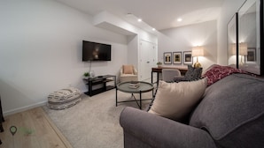 Un salon moderne avec un canapé gris, une petite table basse ronde, un téléviseur fixé au mur et une porte blanche en arrière-plan.