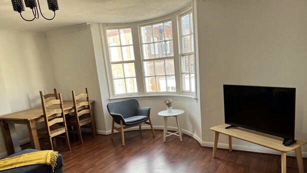 A living room with a wooden dining table, chairs, a sofa, a television on a stand, and a window with a view of buildings. - Spacious 1-bed - Coolest Neighbourhood in Brighton (Brighton)