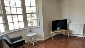 A living room with a television on a stand, a small table with a plant, and a chair. - Spacious 1-bed - Coolest Neighbourhood in Brighton (Brighton)