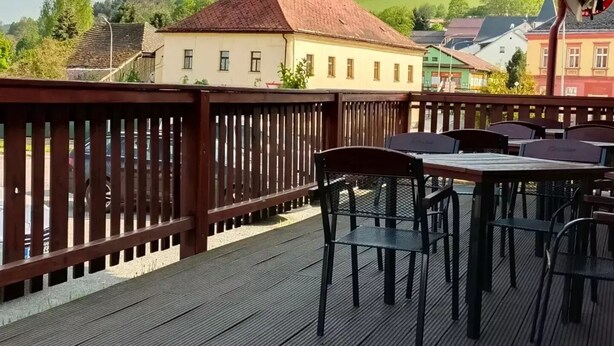 A wooden deck with tables and chairs, overlooking a village with buildings and a green hill in the background. - Hotel Vintíř (Hartmanice)