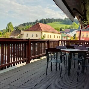 Una terraza de madera con mesas y sillas, con vista a un pueblo con edificios y una colina verde al fondo.