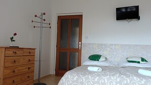 A bedroom with a wooden dresser, a floor lamp, a bed with patterned bedding, a green and white pillow, and a flat-screen TV mounted on the wall. - Hotel Vintíř (Hartmanice)