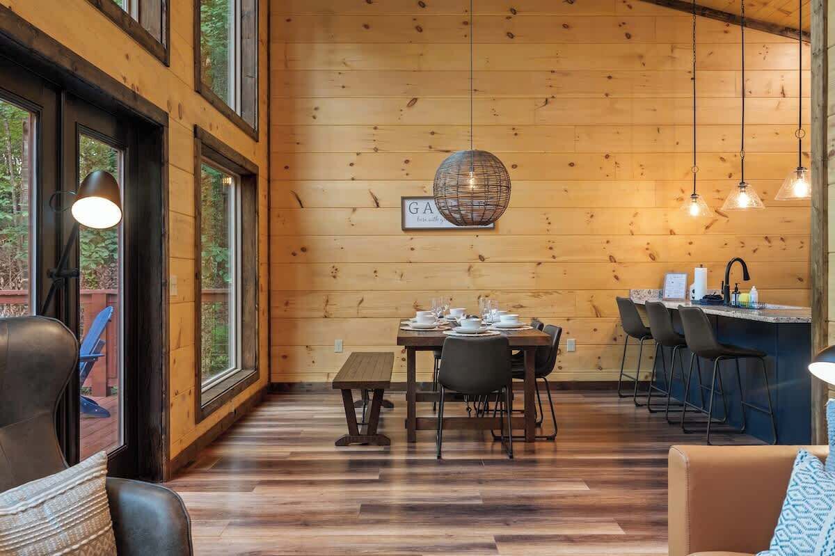 A cozy cabin interior with a wooden dining area, a bar, and large windows.