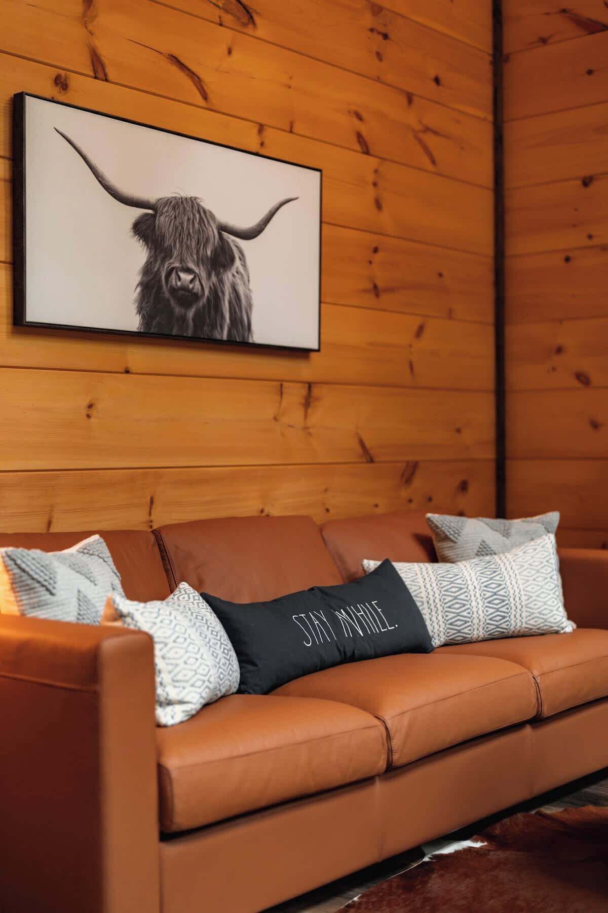 A brown leather sofa with patterned and solid pillows, a framed artwork of a yak, and wooden paneling on the wall.