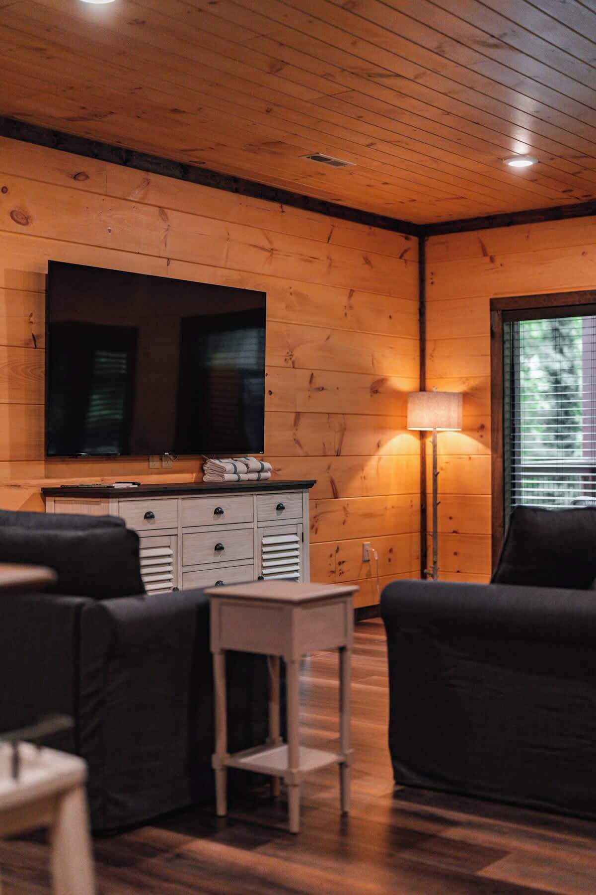 Una habitación acogedora con paredes de madera, un televisor de pantalla plana montado sobre un mueble de madera, una mesita y dos sofás negros.