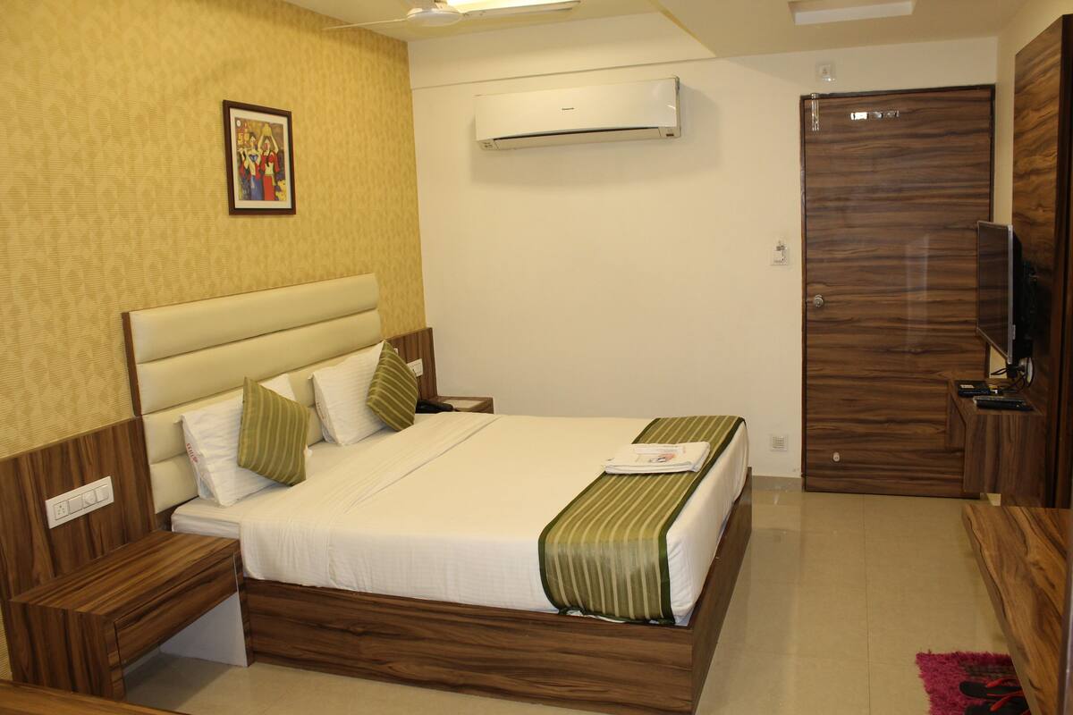 A hotel room with a bed, bedside tables, a wardrobe, an air conditioning unit, and a television.