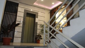 Una entrada de edificio moderno con un muro de mármol, un ascensor verde y una escalera con barandillas blancas.