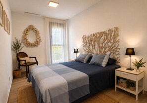 Et soveværelse med seng, stol, natbord, lampe, billedkunst på væggen og et vindue med gardiner.