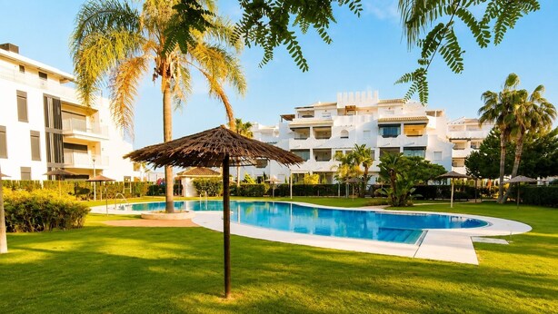 A swimming pool surrounded by a well-maintained lawn, palm trees, and modern buildings. - Rota Punta Candor Terrace Playa 3 Bajo C (AN)