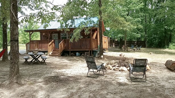 A rustic cabin with a wooden deck, picnic table, and two chairs in a wooded area.