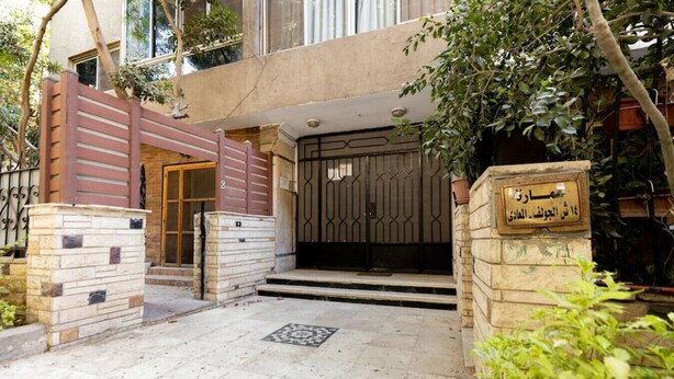 A gated entrance with a stone pillar and a metal gate, surrounded by greenery and a paved area. - Lily Mint Comfy 3BR W/Sitting room TV Apartment (Maadi as Sarayat Al Gharbeyah)