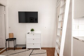 Una habitación blanca con una televisión montada en la pared, un tocador blanco con cajones, una planta pequeña encima y una mesita de noche negra, además de una alfombra blanca.