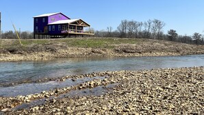 A two-story house with a large deck, situated by a river with a rocky shoreline.