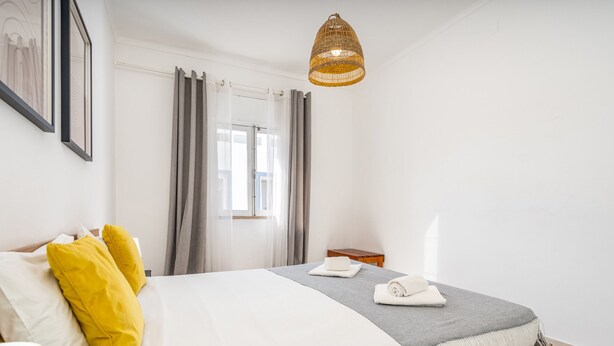 Una habitación de hotel con cama, ventana con cortinas, mesita de noche y lámpara colgante.