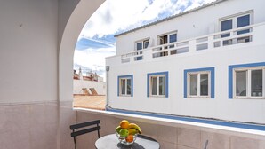 Un balcón con una mesa, un frutero y una silla. Se ve un edificio blanco con detalles azules y ventanas al fondo.