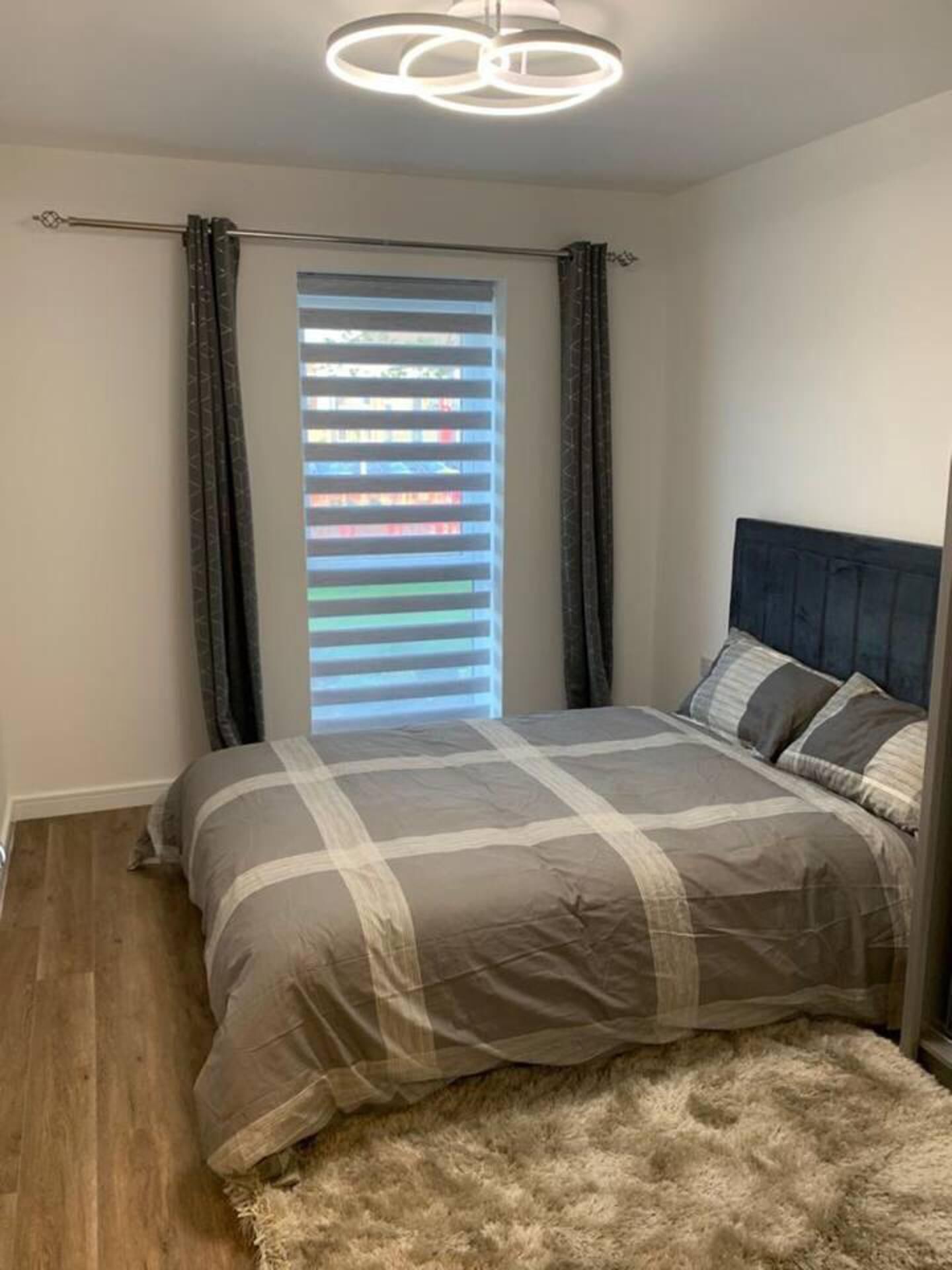 A bedroom with a bed, a headboard, a window with blinds, and a ceiling light fixture.