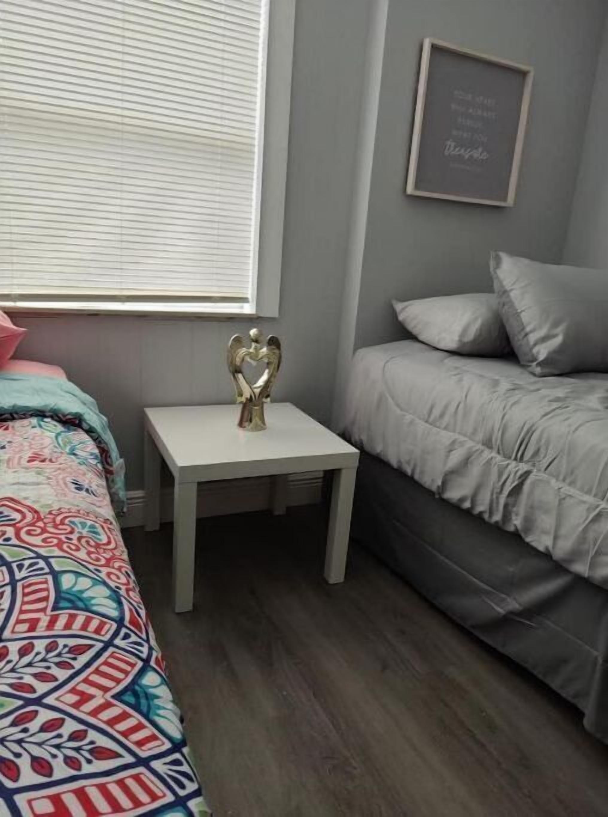 A bedroom with a bed, a bedside table with a decorative item, a window with blinds, and a framed quote on the wall.