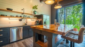 A modern kitchen with a wooden countertop, stainless steel appliances, and a large window overlooking a forest.