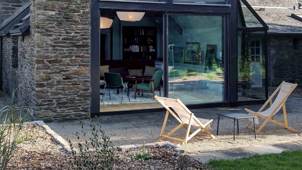 A modern house with a stone chimney, large glass windows, and a cozy interior with a dining area and seating. - La Maison de L'amiral (Moëlan-sur-Mer)