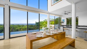 A modern kitchen with a large island, a dining area with a wooden table set for six, and a view of the ocean through large windows. - Stunning contemporary villa with incredible views over the Barbados West Coast (Lower Carlton)
