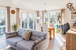 A modern living room with a grey sofa, a dining table with chairs, large windows, and a view of the outdoors. - St Helens 2-bed Lodge, Sleeps 4, Pets Ok & Pool (Ryde)