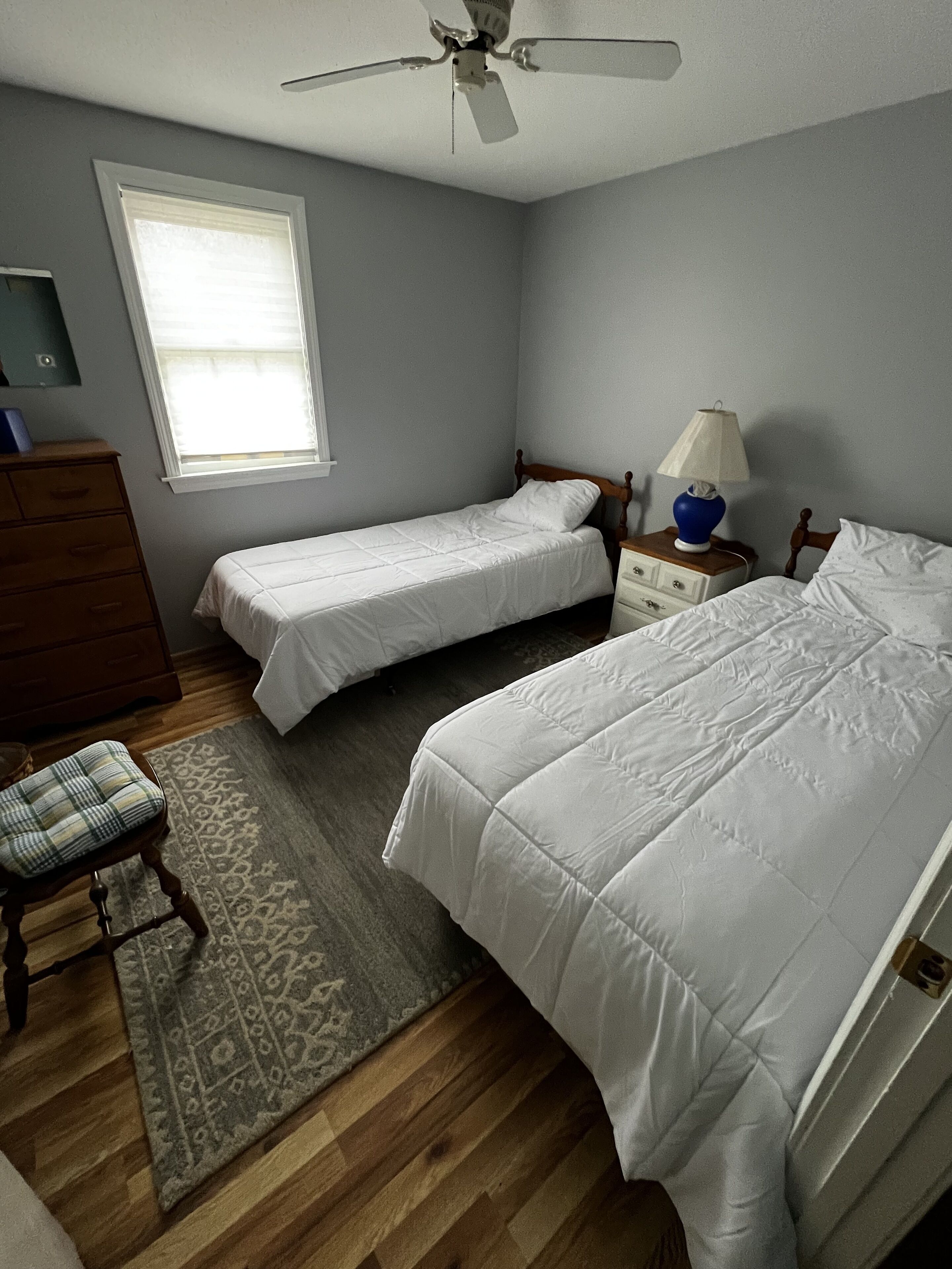 Ein Schlafzimmer mit zwei Einzelbetten, einer Holzkommode, einem Nachttisch mit Lampe, einem Deckenventilator und einem Fenster mit Jalousien.