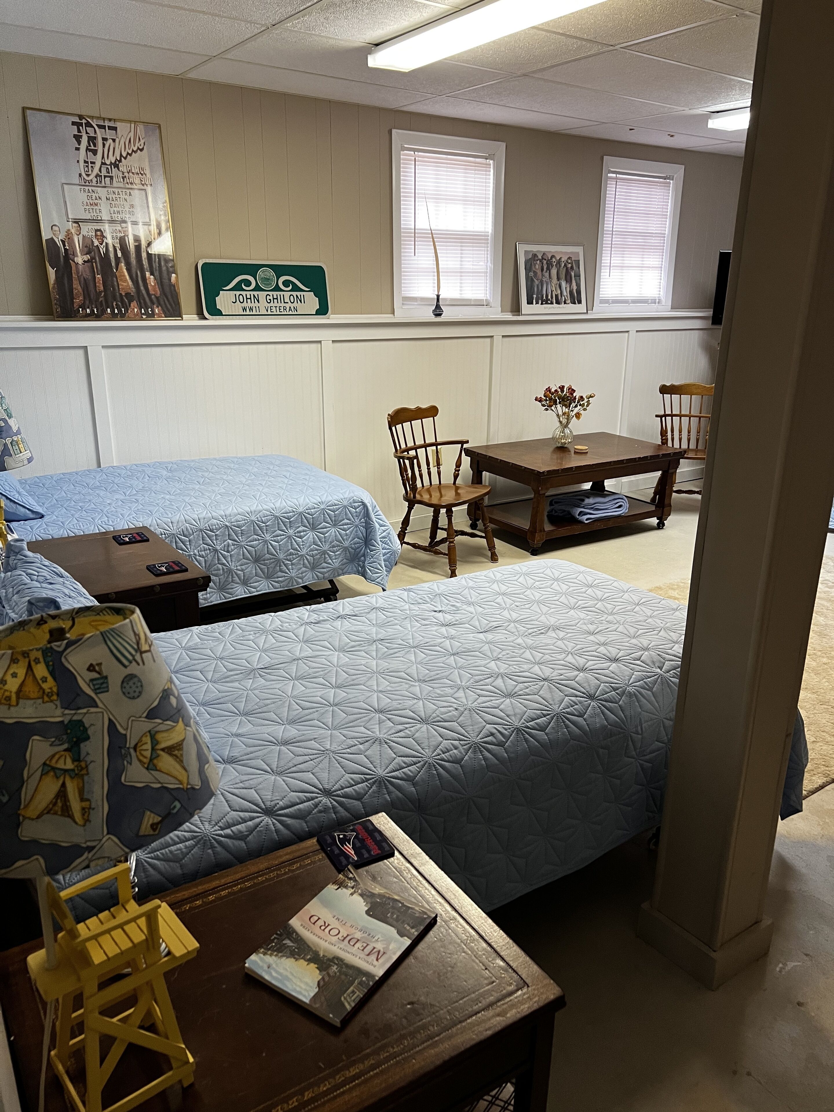A hotel room with two beds, a wooden table with a vase, a book, and a lamp. There are framed pictures on the wall and a sign about a WWII veteran.