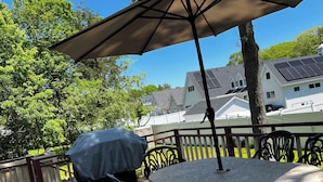 Um deck de madeira com mesa redonda e cadeiras, churrasqueira coberta e um grande guarda-sol.