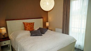 Cama bem arrumada com edredom branco, duas almofadas e cabeceira de madeira. Tem uma mesa de cabeceira com abajur e um objeto decorativo pequeno. Uma luminária redonda e texturizada pendurada no teto.